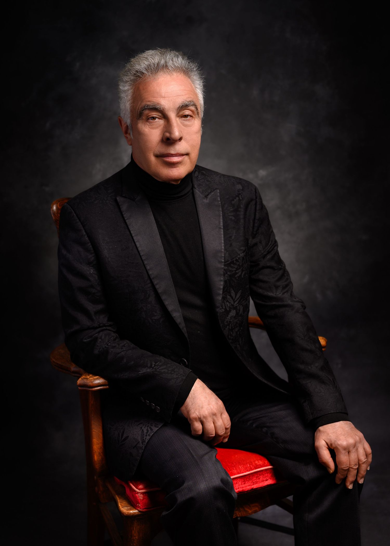 Elegant man in black suit and turtleneck seated in ornate wooden chair with red cushion against dark background.