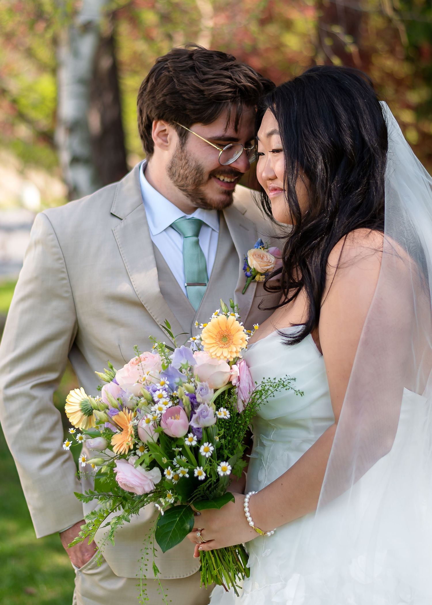 Bride and groom smiling together outdoors, bride holding pastel spring bouquet with roses and daisies, groom in tan suit with mint tie.