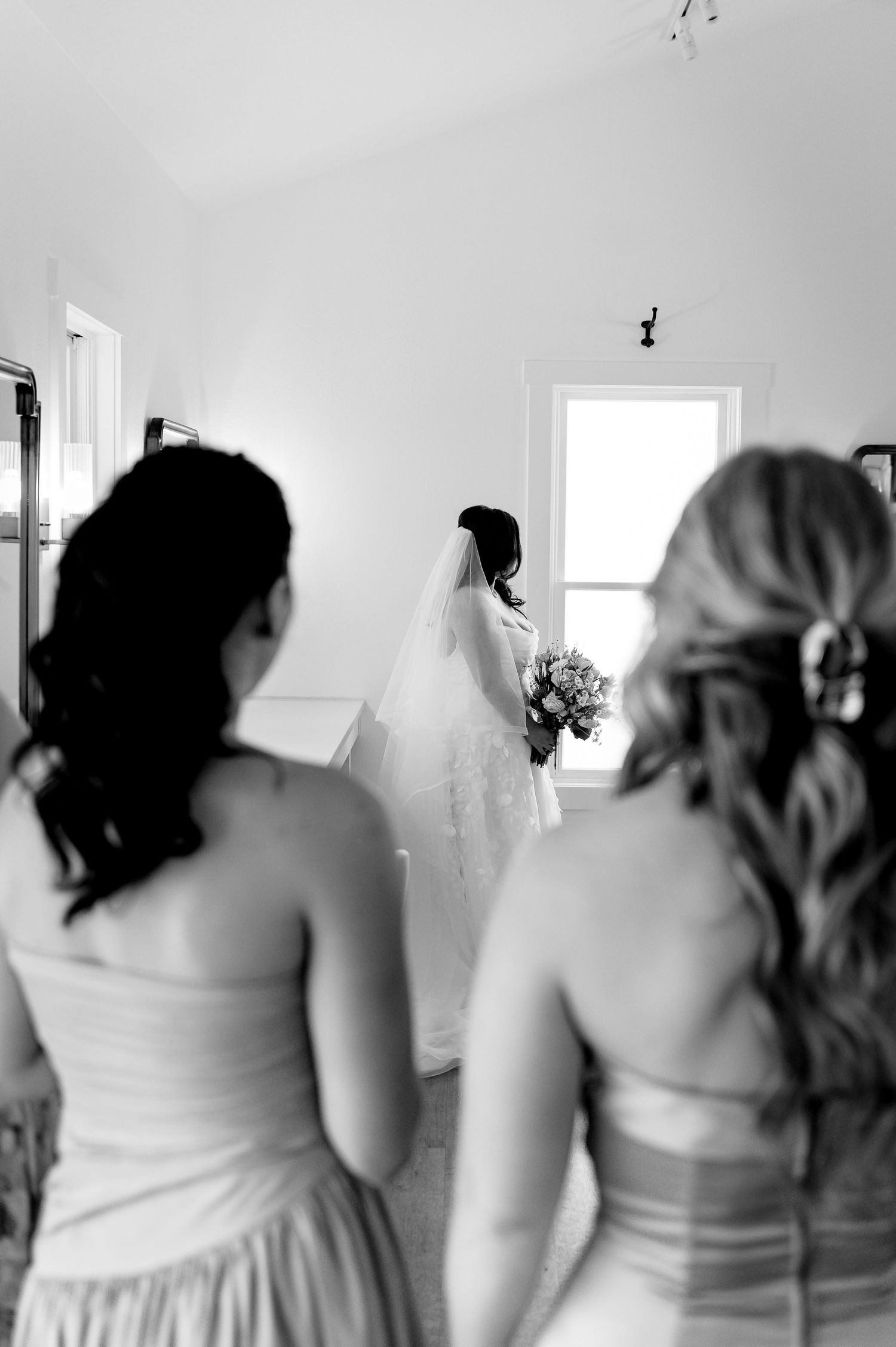 Black and white photo of a bride in veil holding bouquet by window, watched by two bridesmaids from behind.