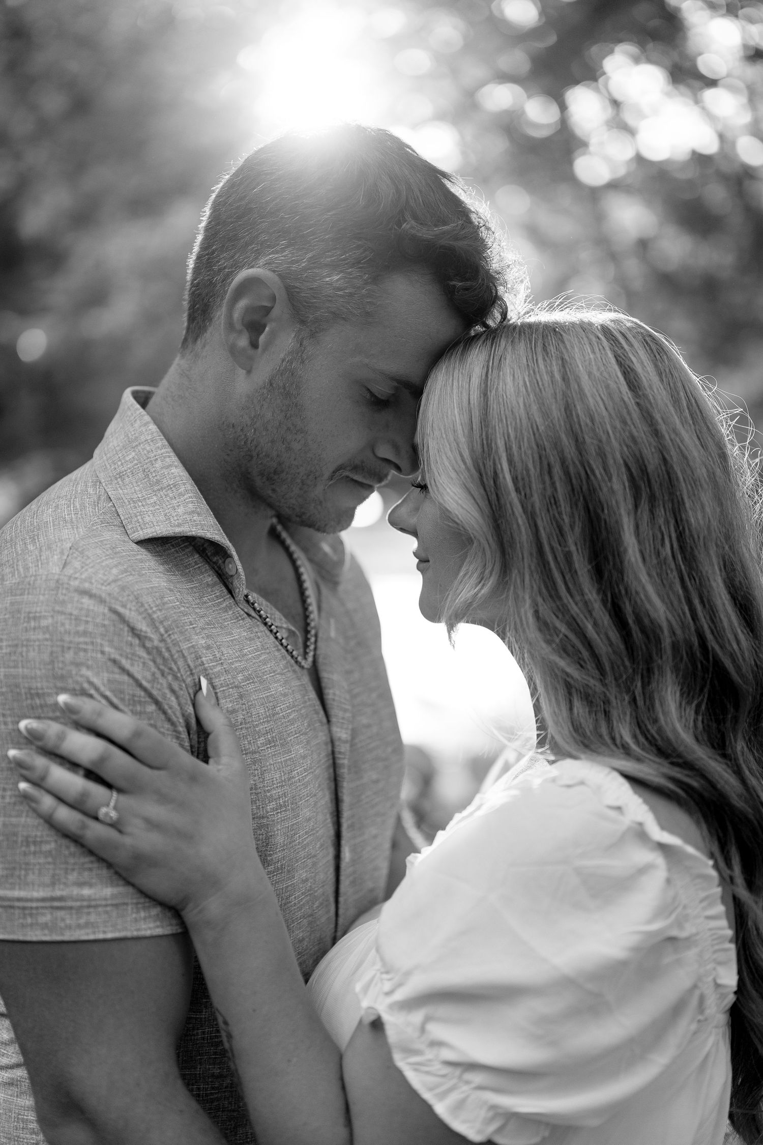 Black and white photo of a romantic couple touching foreheads, embracing outdoors with soft bokeh sunlight behind them.