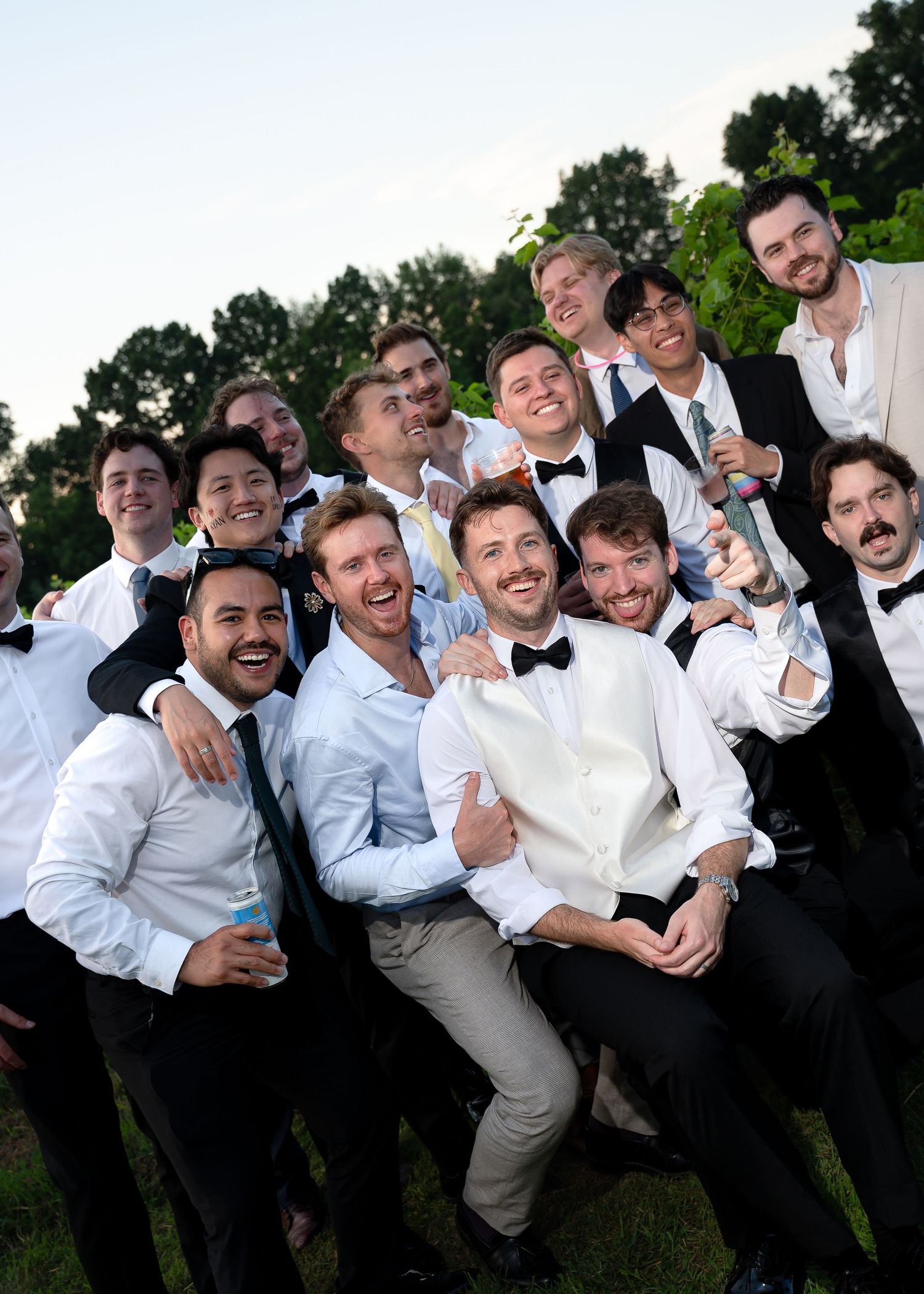 Group of men in formal tuxedos and suits smiling and posing together outdoors at a wedding celebration.