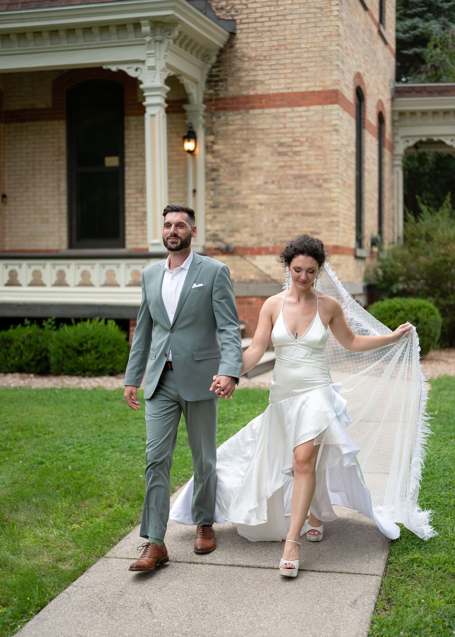 Bride in white gown holding veil and groom in gray suit walk hand in hand on sidewalk outside brick Victorian building.