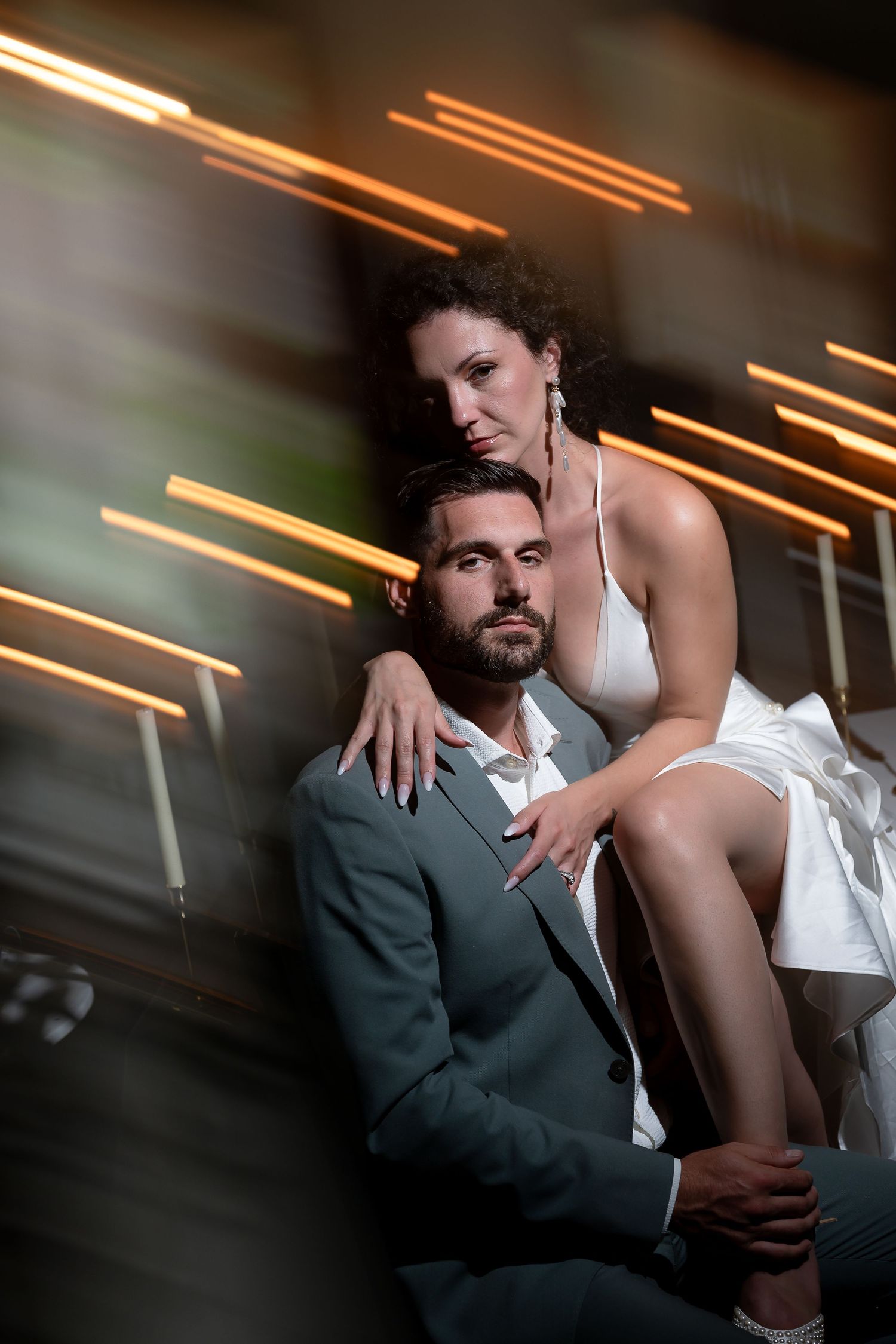 Elegant couple posing together, woman in white dress embracing man in gray suit, with dramatic light streaks in background.