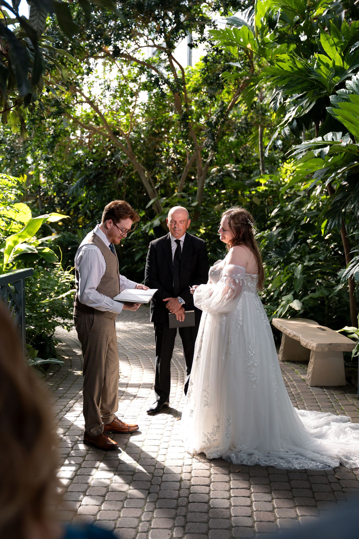 Couple exchanges vows in lush garden wedding ceremony with officiant, bride in lace gown, groom in brown suit.