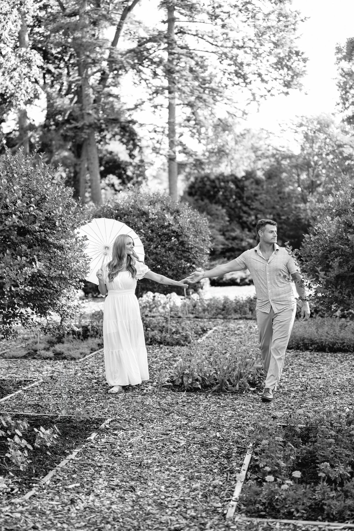 Black and white photo of a couple holding hands walking through a lush garden, woman in white dress holding parasol.