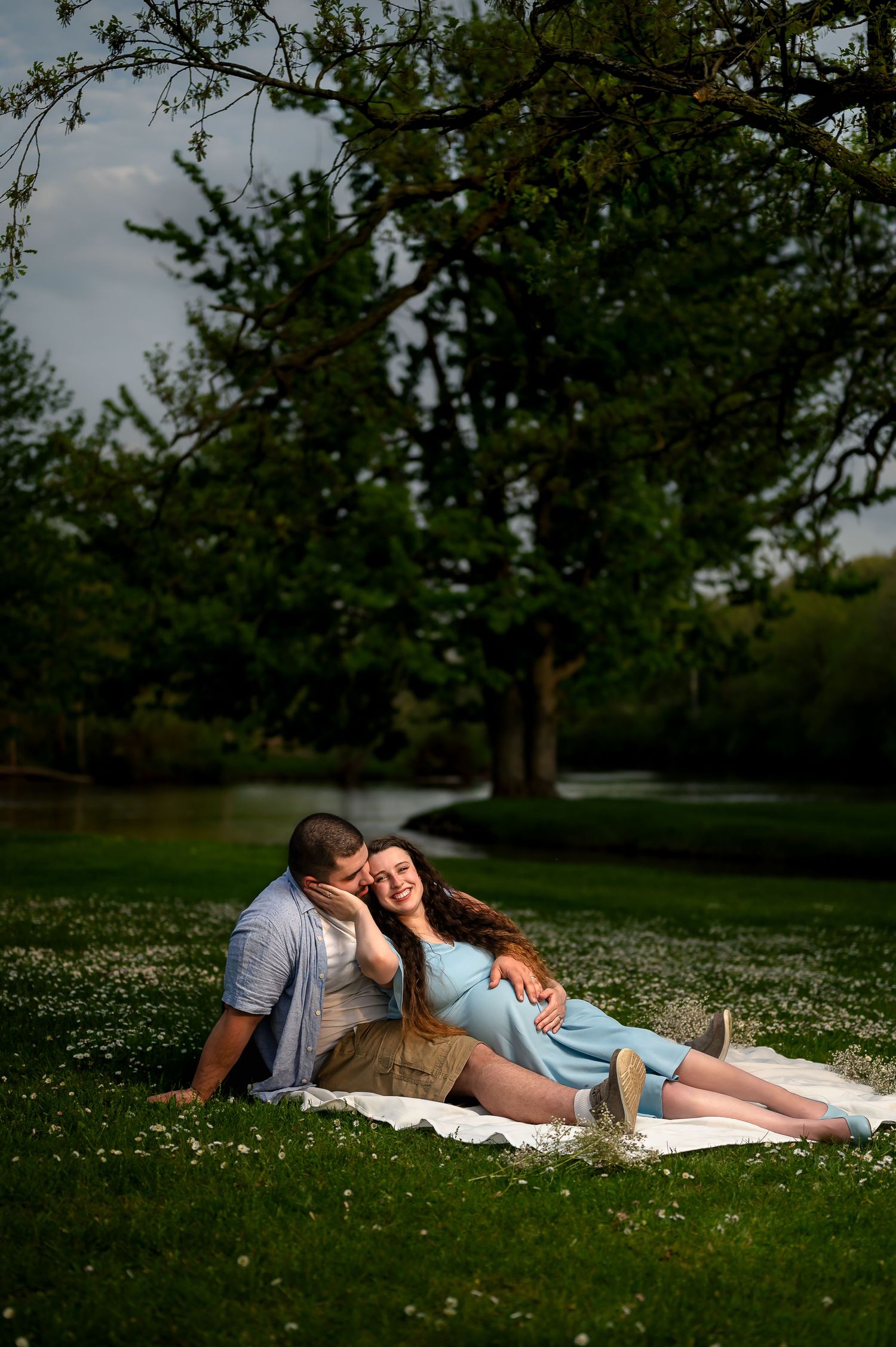 Pregnant couple relaxing on a blanket in a lush green park, man tenderly embracing smiling woman in blue maternity dress.
