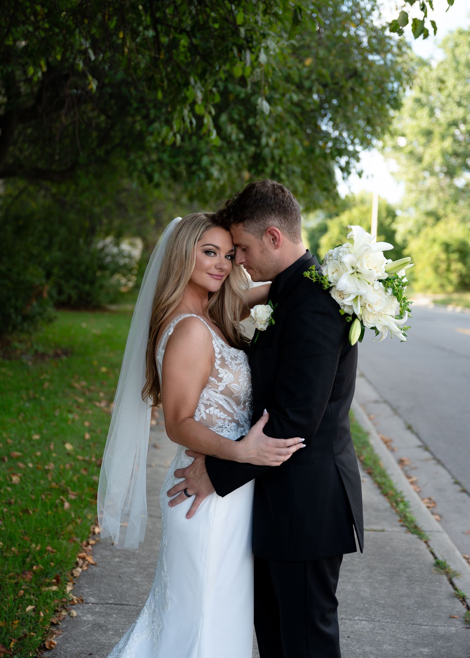 Bride in white lace gown and veil holds white floral bouquet while embracing groom in black suit outdoors.