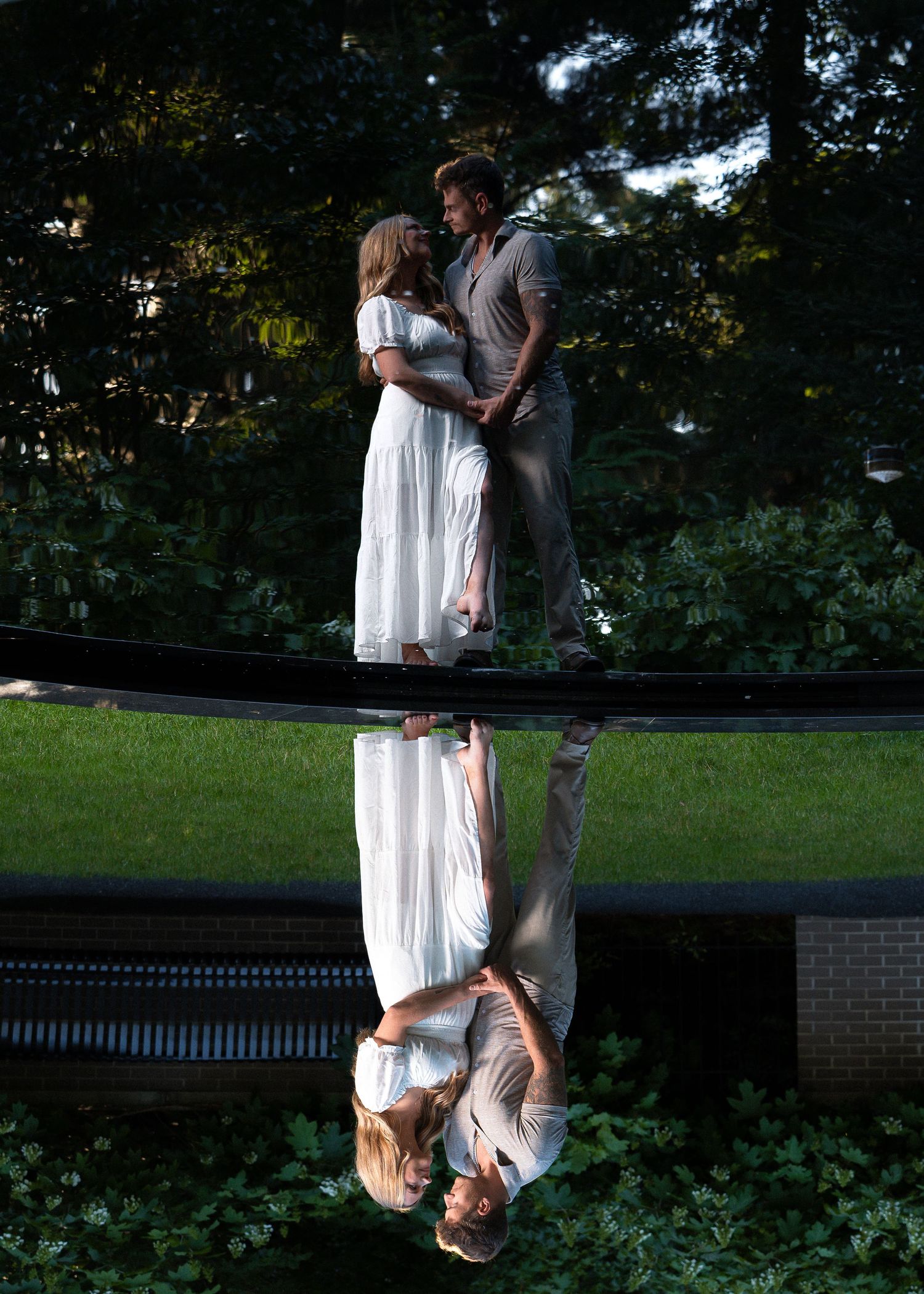 Couple in white dress and gray outfit embracing near reflective pool with lush green trees backdrop.
