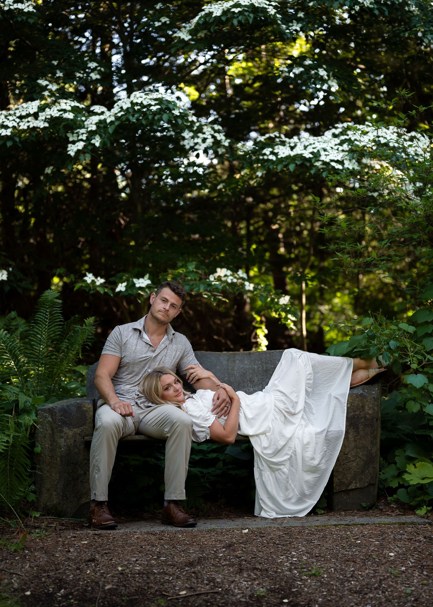 Couple relaxing on a stone bench surrounded by lush greenery, woman in white dress resting on mans lap outdoors.