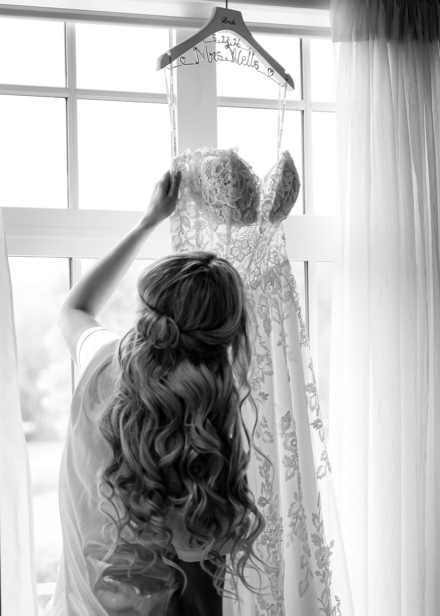 Bride with curly hair admiring her lace wedding dress hanging by a window in a black and white photo.