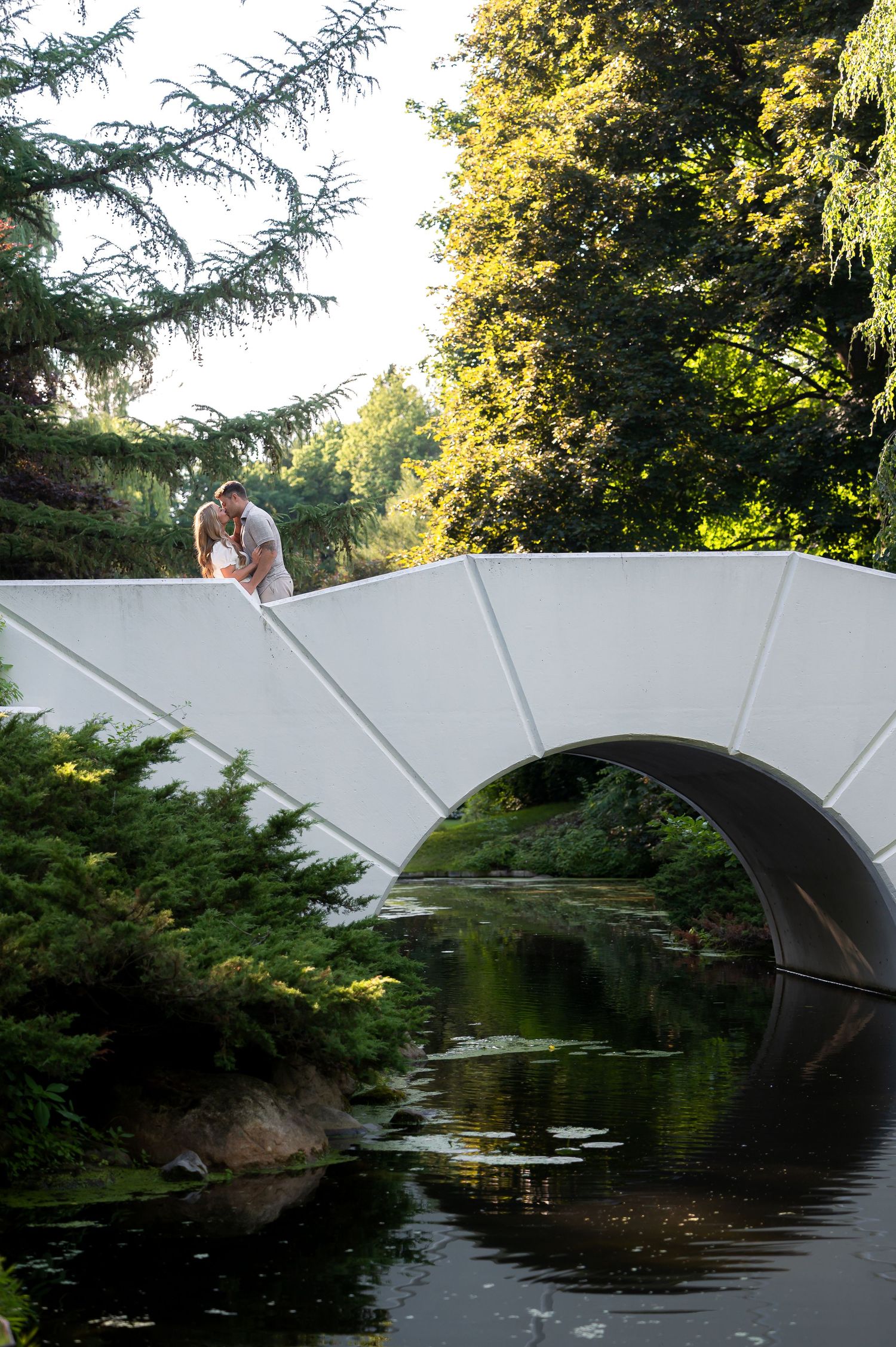 Couple sharing a romantic kiss on a white arched bridge surrounded by lush green trees reflected in calm water below.