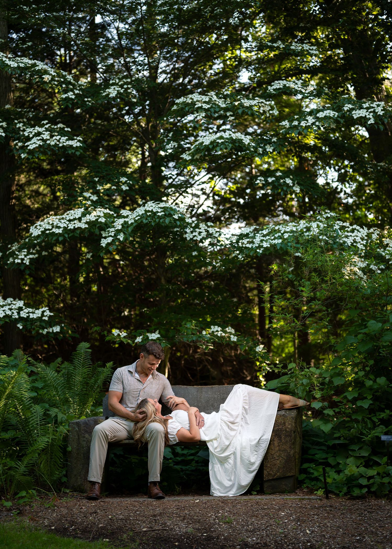 Couple sharing a romantic moment on a stone bench surrounded by lush green trees and white flowering shrubs in a garden.