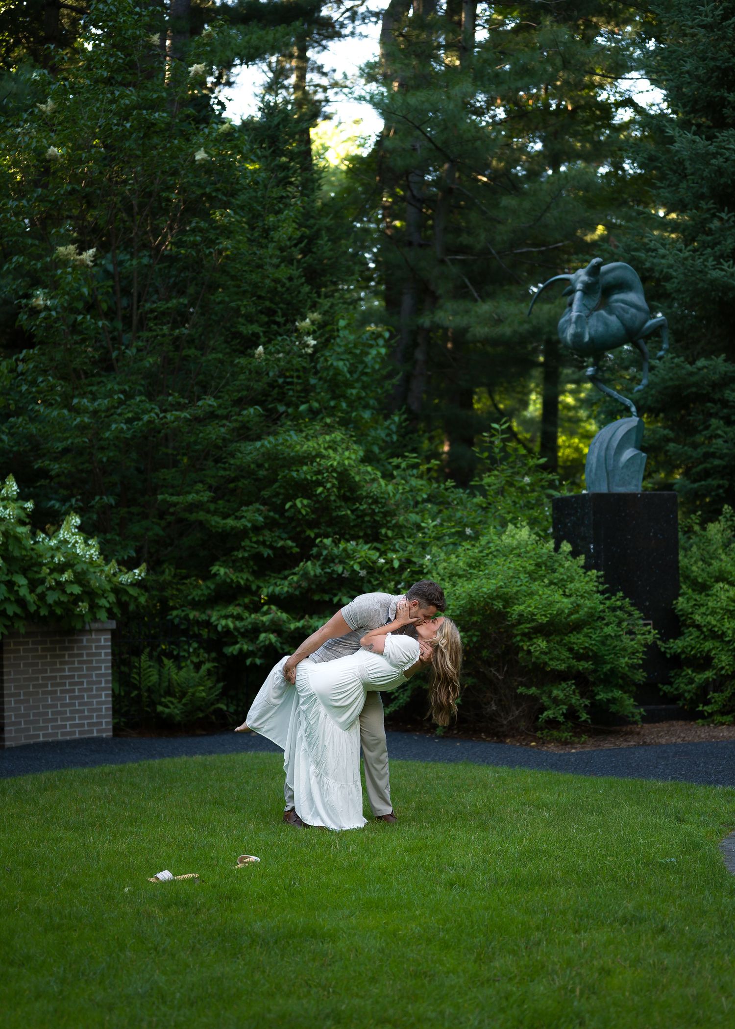 Couple sharing a romantic dip kiss on lush green lawn with garden statuary and trees in background.