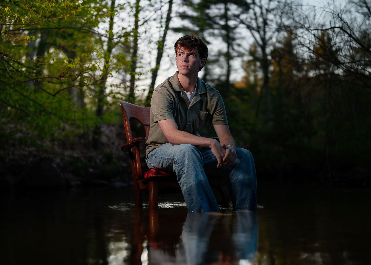 Young man sitting on a wooden chair in a flooded forest stream, surrounded by trees in soft evening light.