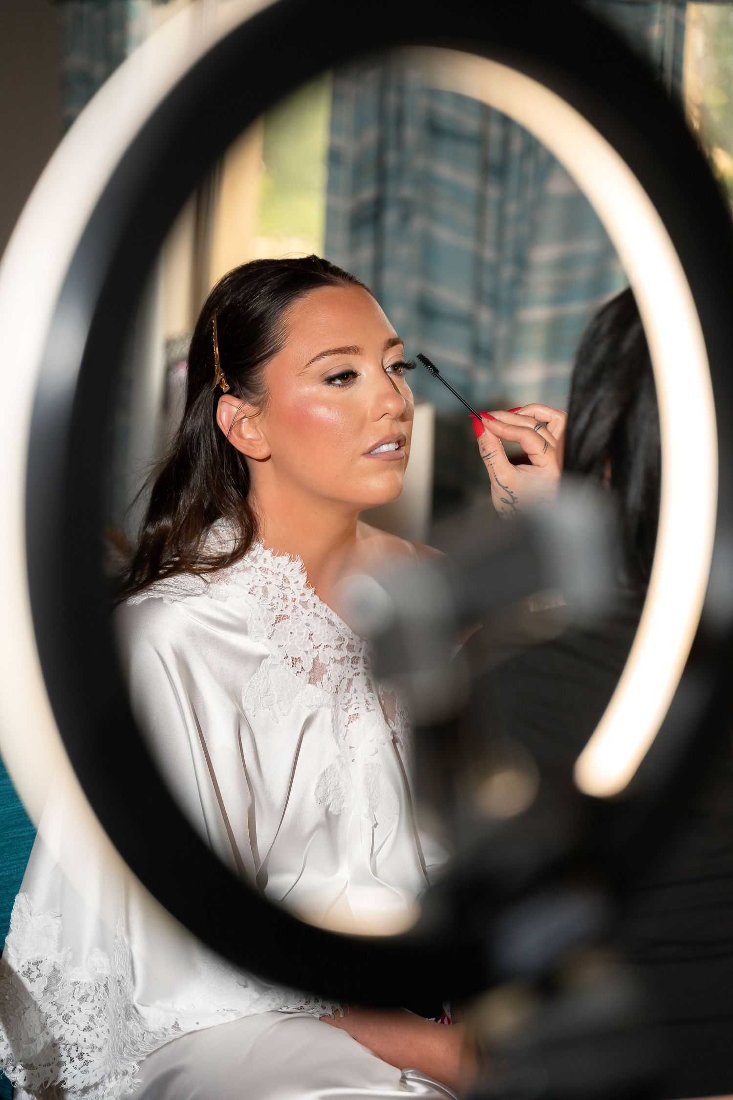 Bride in white lace robe gets makeup applied by artist, framed through a ring light during bridal preparations.