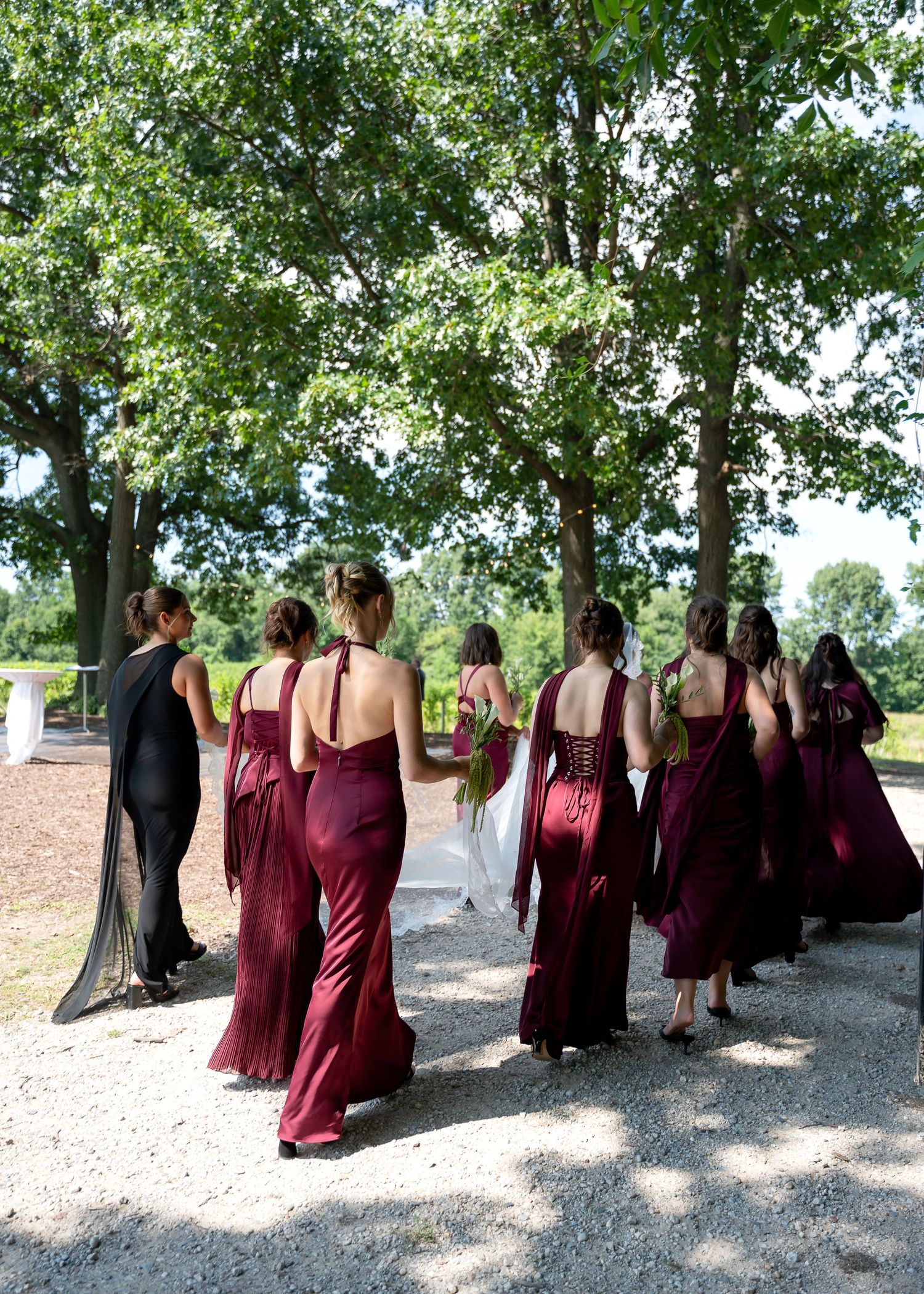Bridesmaids in burgundy gowns walk with bride in white dress through tree-lined path at outdoor wedding.