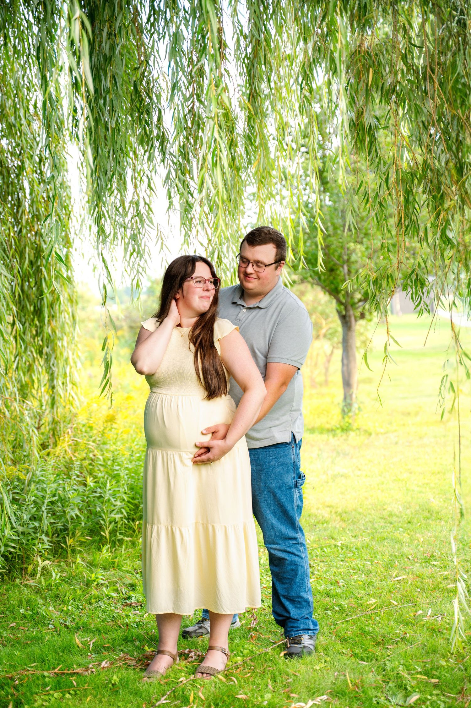 Romantic couple embracing under weeping willow trees in sunlit park setting.