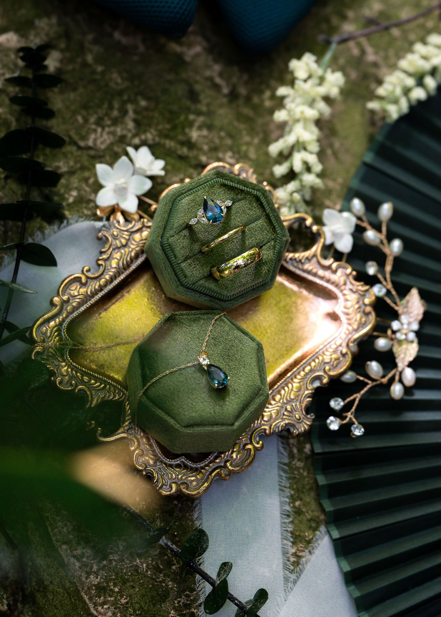 Elegant green velvet ring boxes on gold tray with blue gemstone rings, necklace, and white floral accents.
