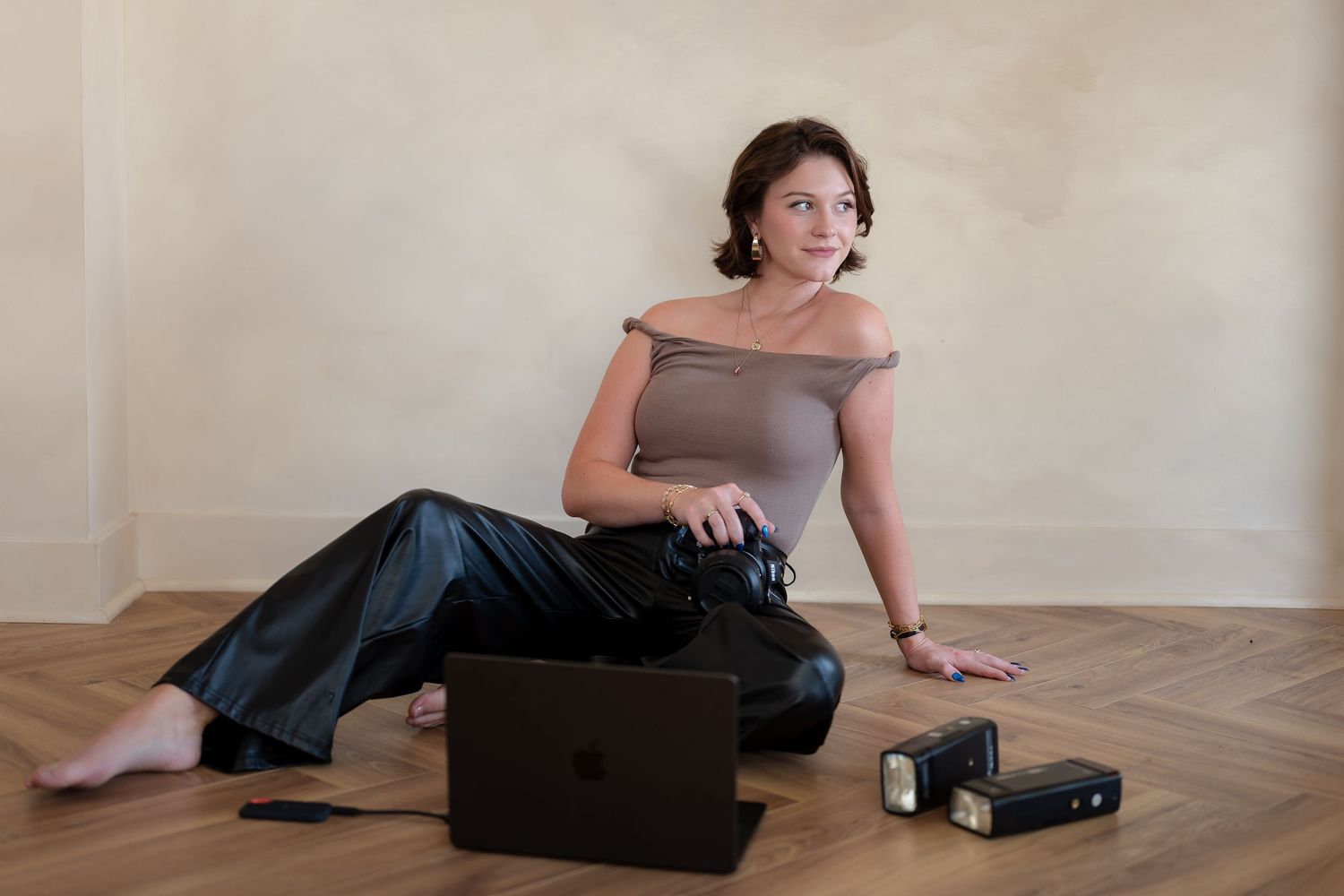 Woman sitting on wooden floor with laptop and camera equipment, wearing gray off-shoulder top and black satin pants.