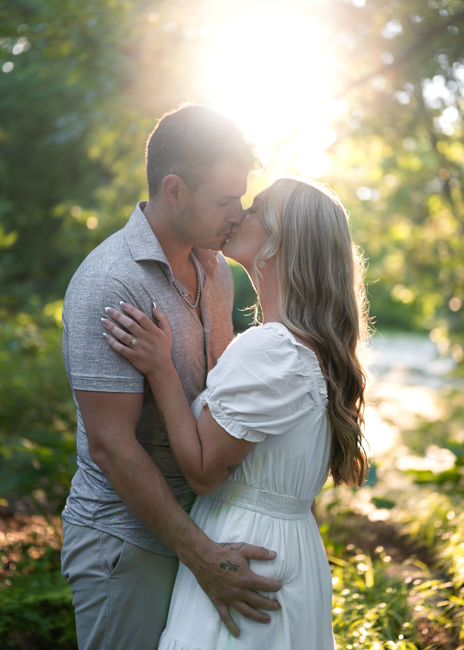 Couple sharing a romantic kiss outdoors in golden sunlight, surrounded by lush green trees near a serene waterway.