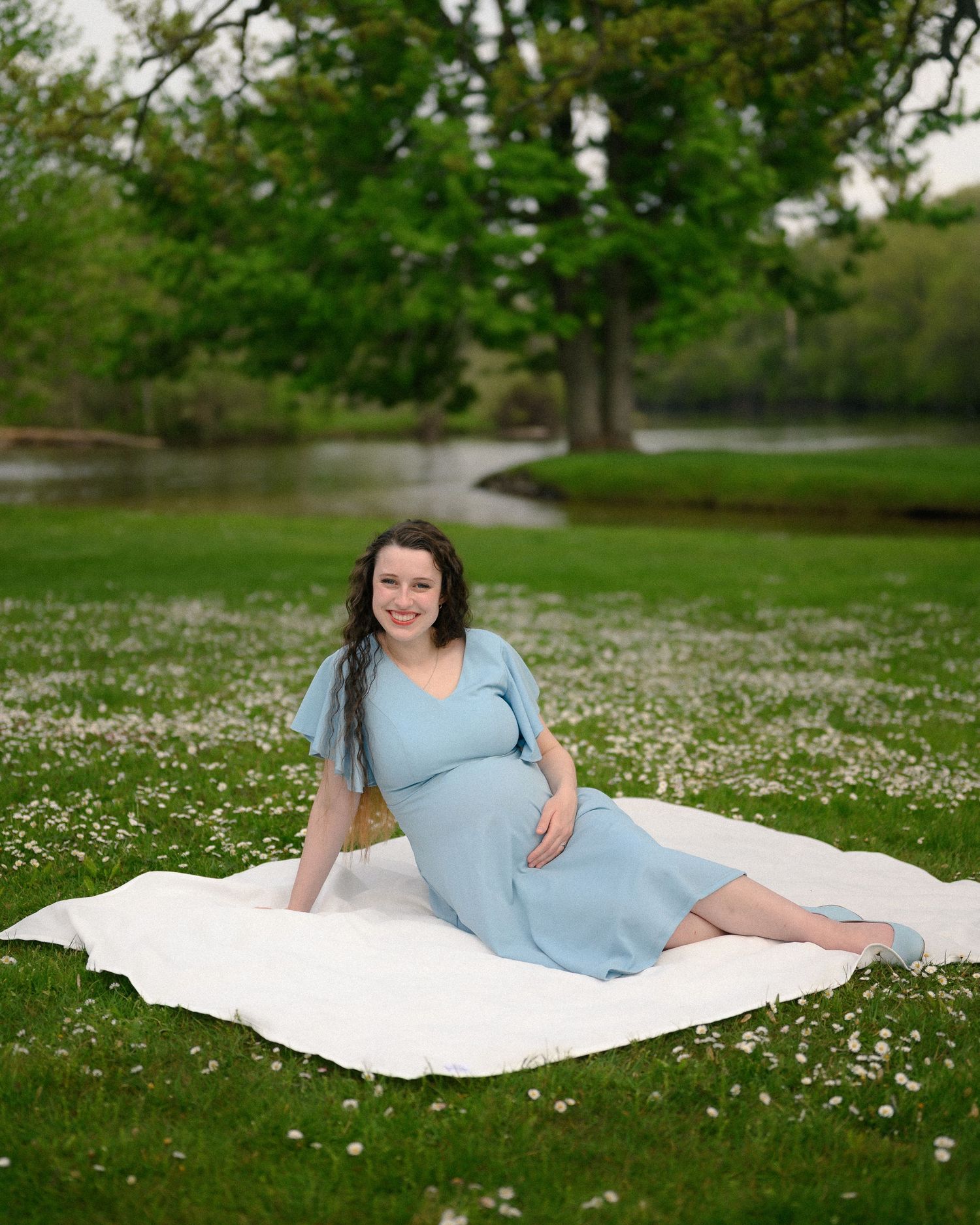 Pregnant woman in light blue dress sitting on white blanket in flower-filled park near a pond.