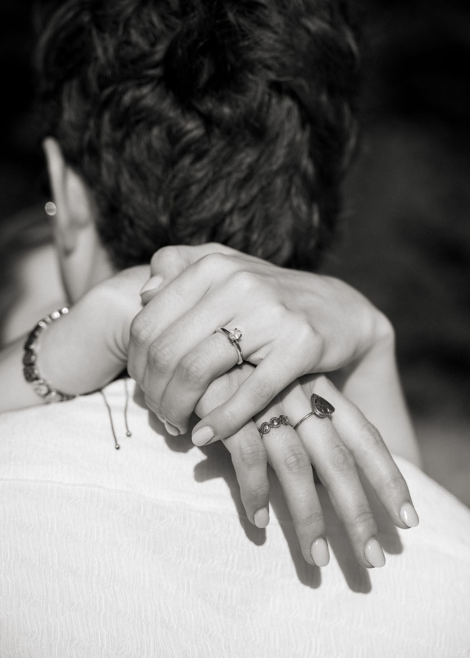 Close-up black and white shot of hands with rings embracing from behind, showing intimate details and jewelry.