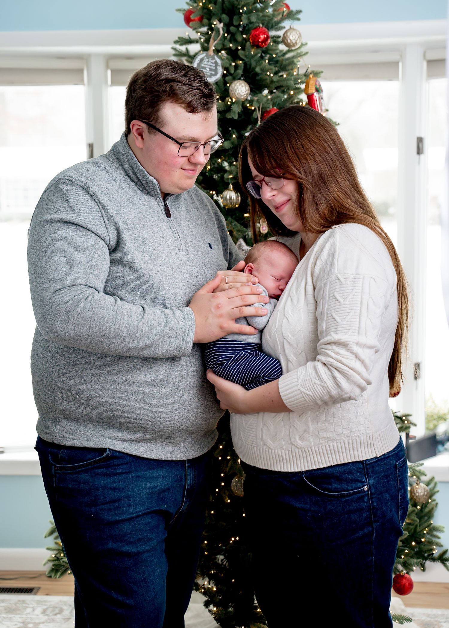A family poses with their baby in front of a decorated Christmas tree in a bright room.