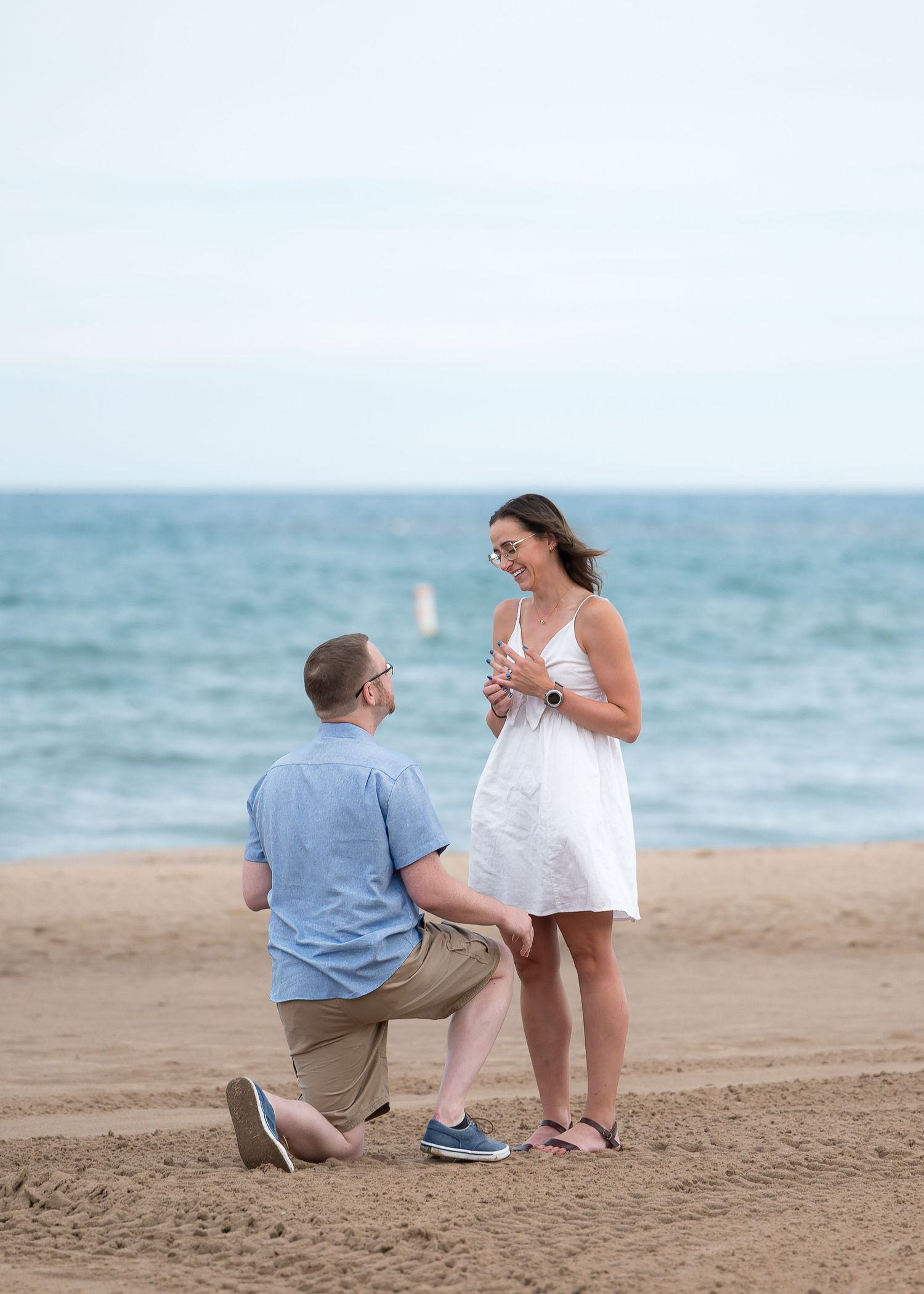 Man kneeling on sandy beach proposing to woman in white dress with ocean in background.