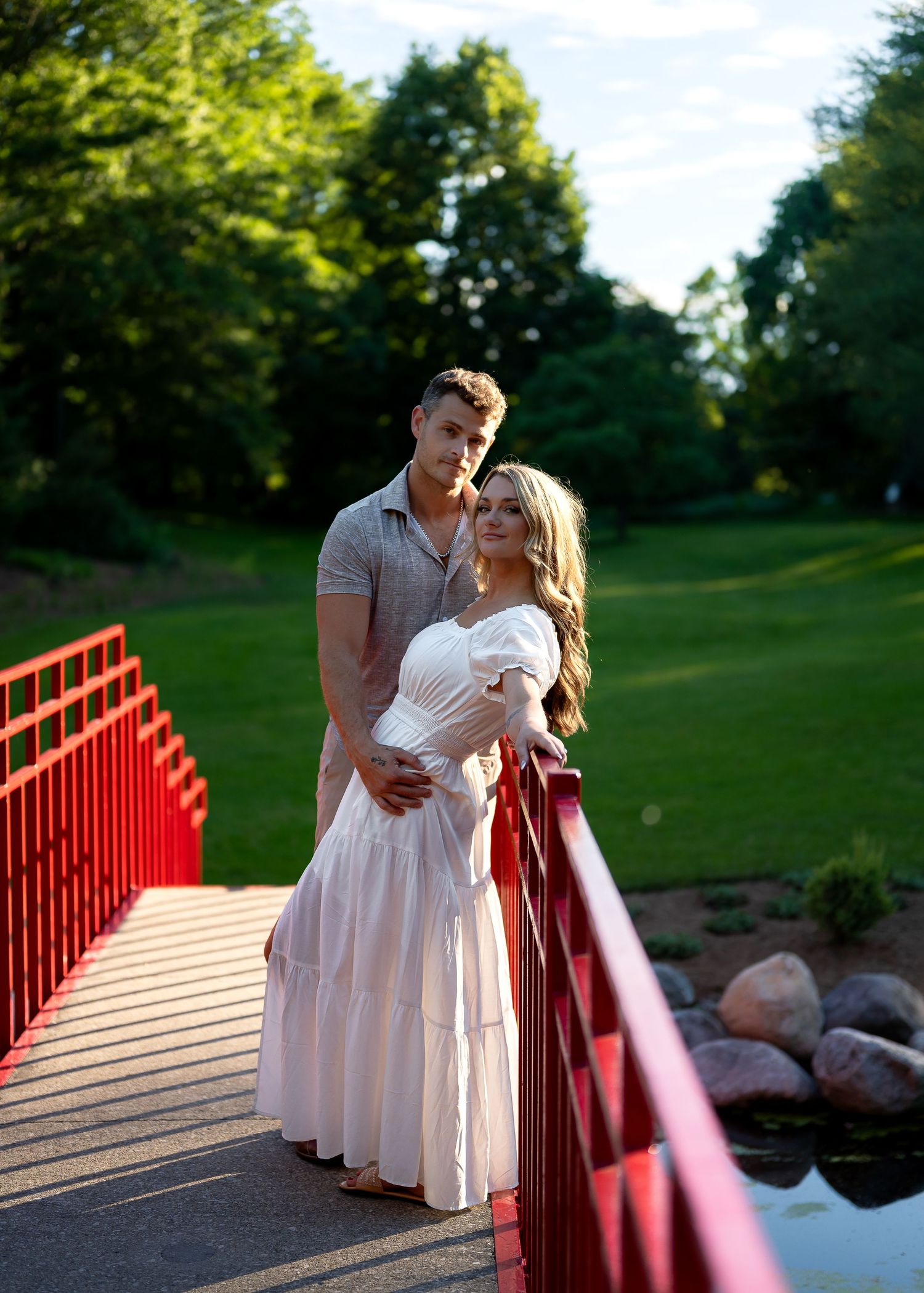 Couple posing on a red bridge surrounded by lush green trees, woman in white dress, man in gray shirt.