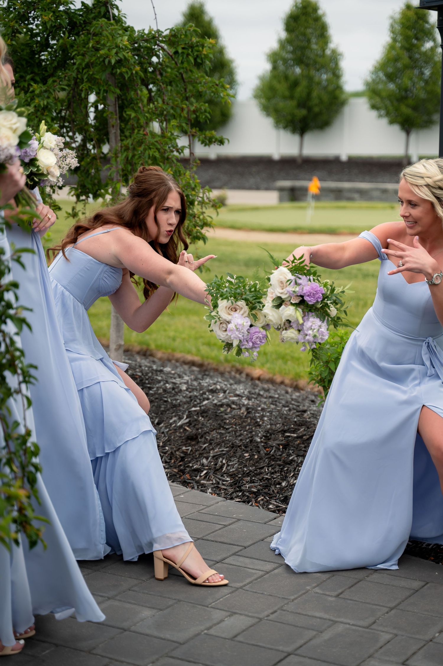 Bridesmaids in light blue gowns playfully pose with floral bouquets outdoors at a wedding venue.