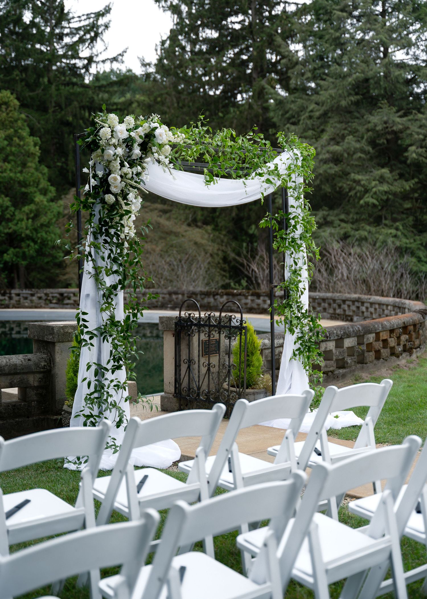 Outdoor wedding ceremony setup with white floral arch, greenery garland, and white folding chairs on a lush green lawn.