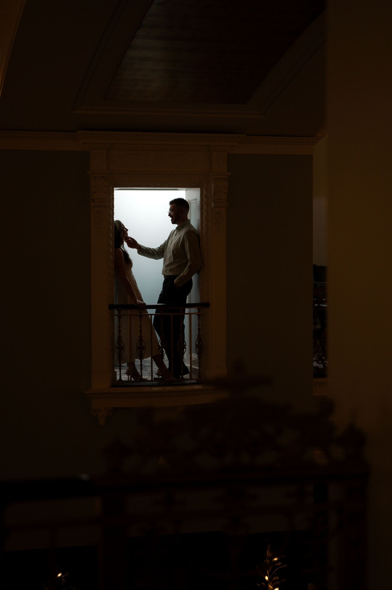 Silhouette of a man standing on a dimly lit balcony at night, framed by a bright window opening.
