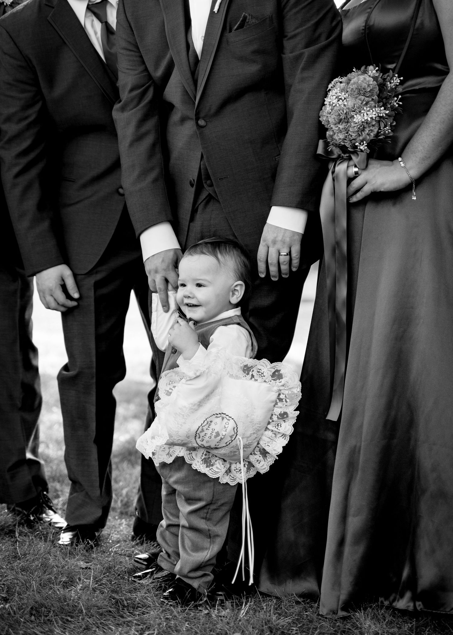 Wedding party members in formal attire stand together while a small child in white ruffles plays at their feet.