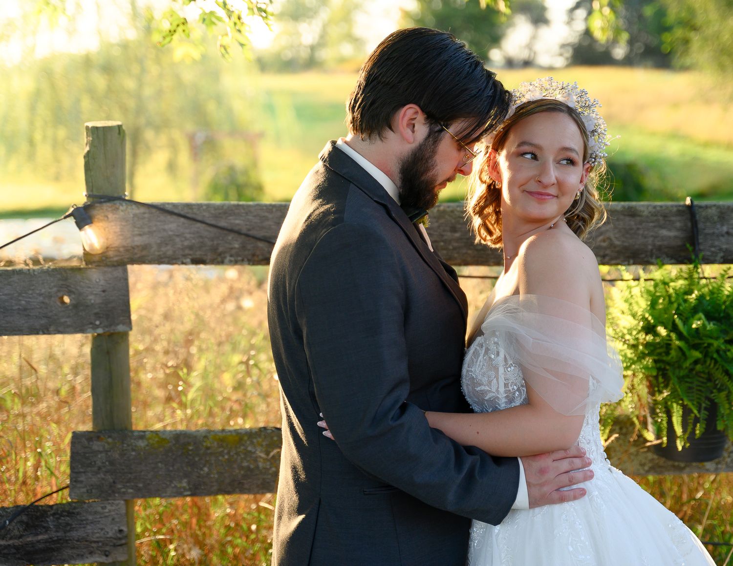 A romantic wedding couple embraces by a rustic wooden fence during golden hour.