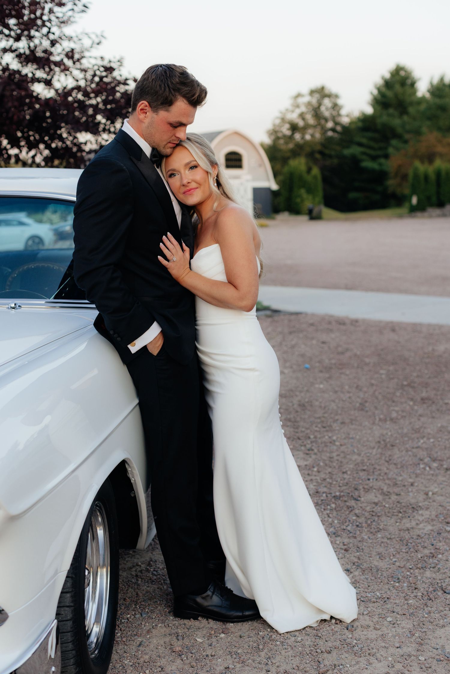 Intimate moment between newlyweds leaning against classic white car during wedding photoshoot.