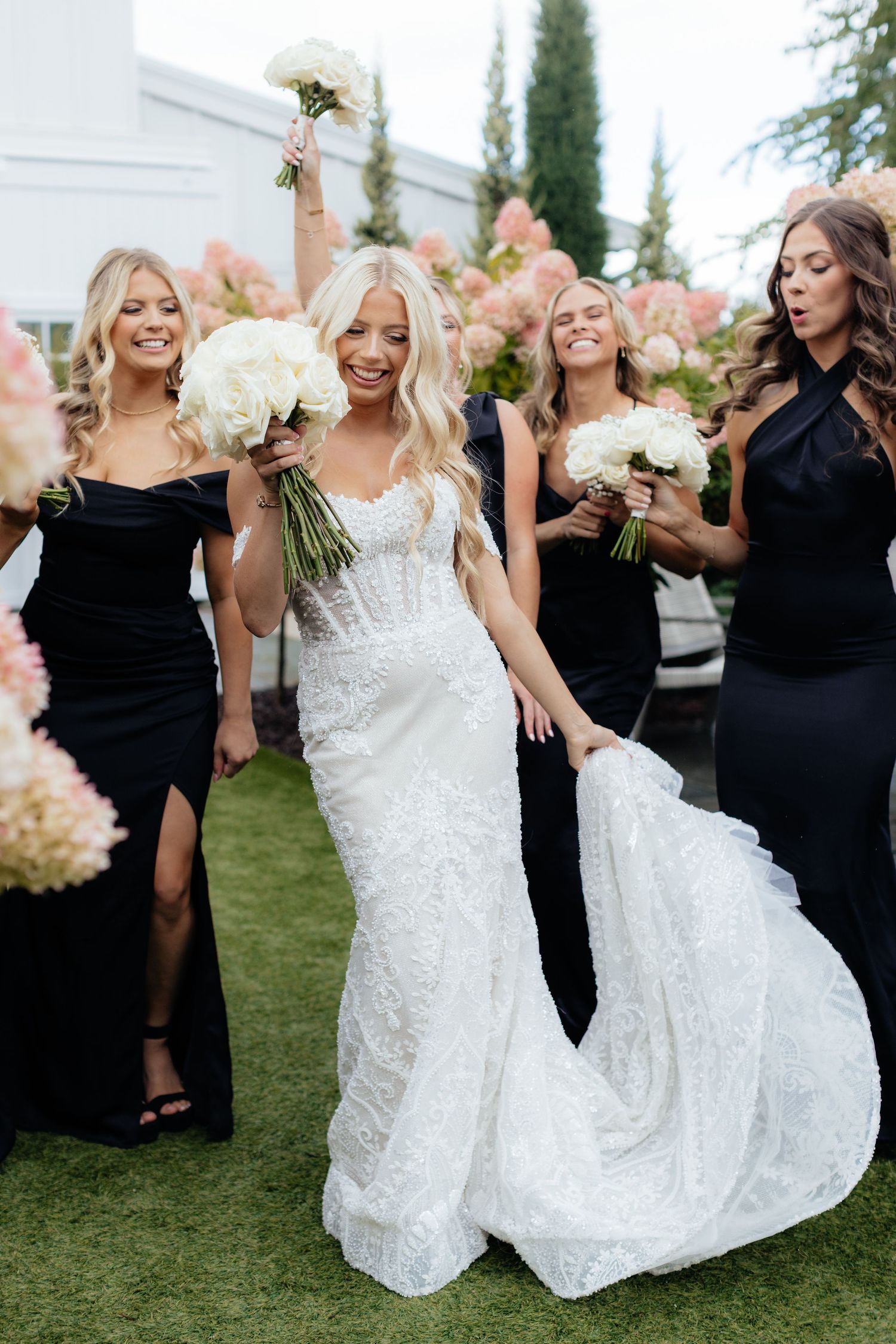 Bridesmaids in elegant black dresses pose with the bride in a lace gown on a manicured lawn.