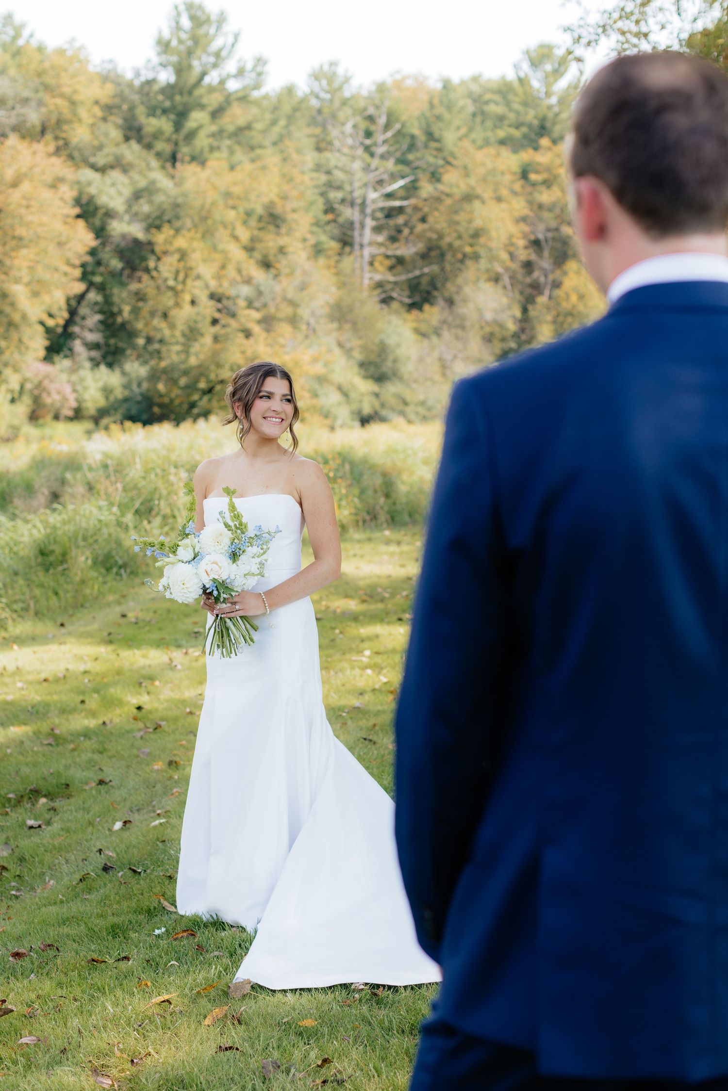 Bride in flowing white dress holds bouquet while posing in grassy outdoor setting.