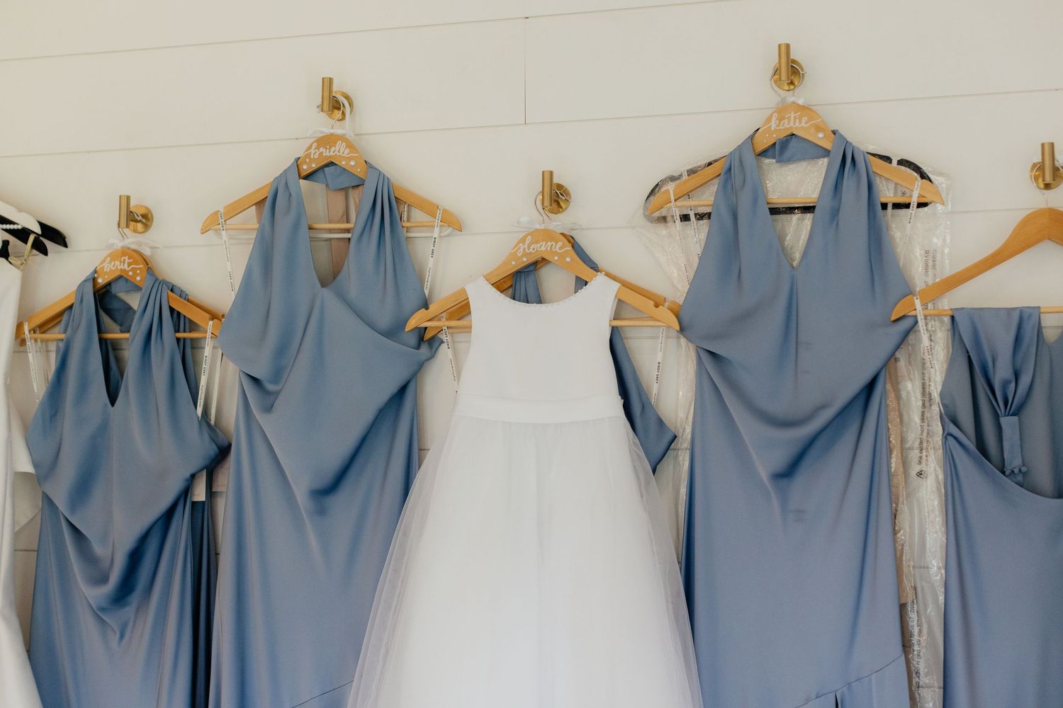 Row of dusty blue bridesmaid dresses hanging on wooden hangers against white wall.