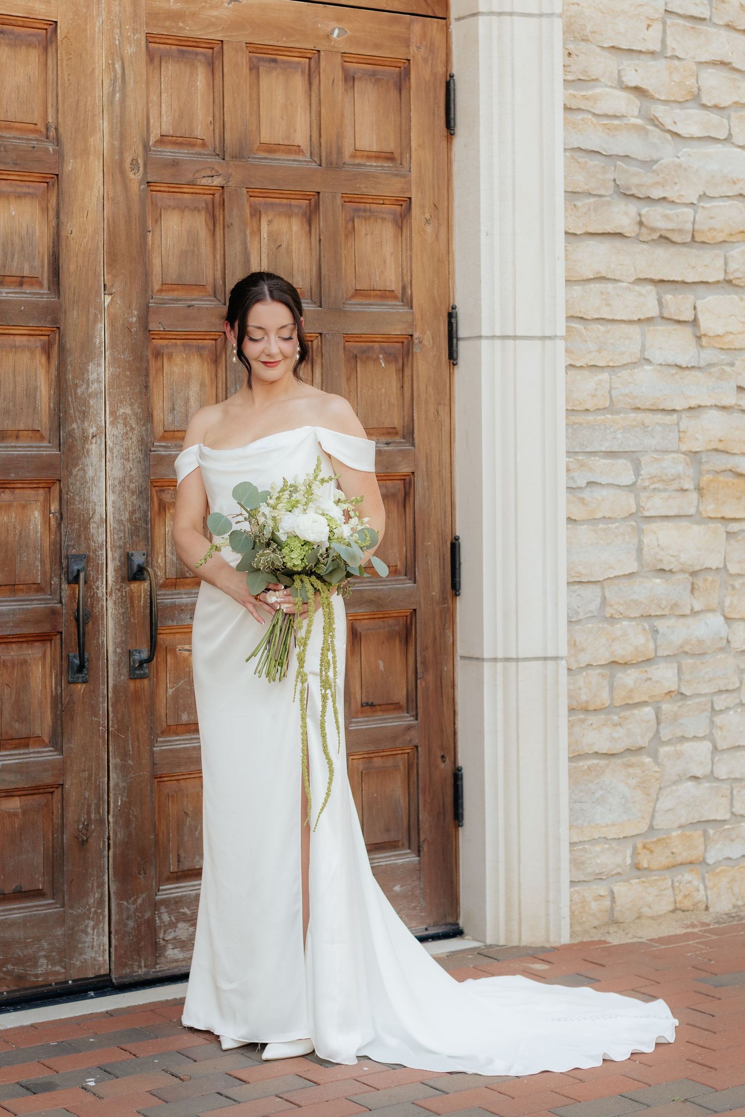 A bride in an elegant off-shoulder white wedding gown poses with a bouquet against rustic wooden doors and stone walls.