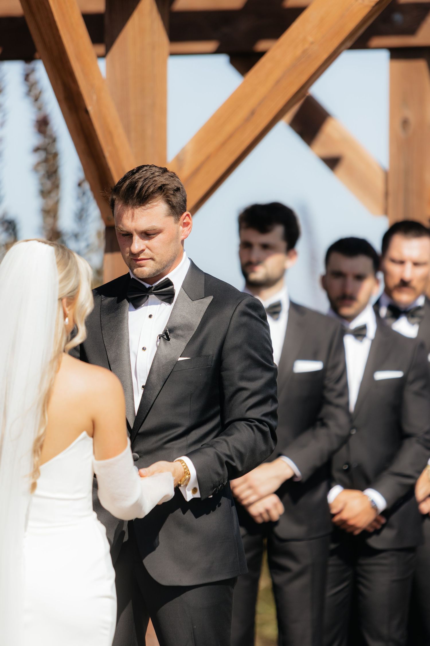 Wedding ceremony taking place under a wooden pergola with guests in formal attire.