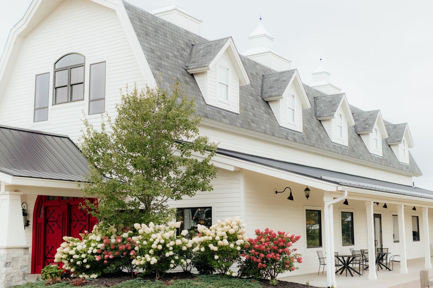 White farmhouse-style venue with dormer windows and flowering bushes along the porch.