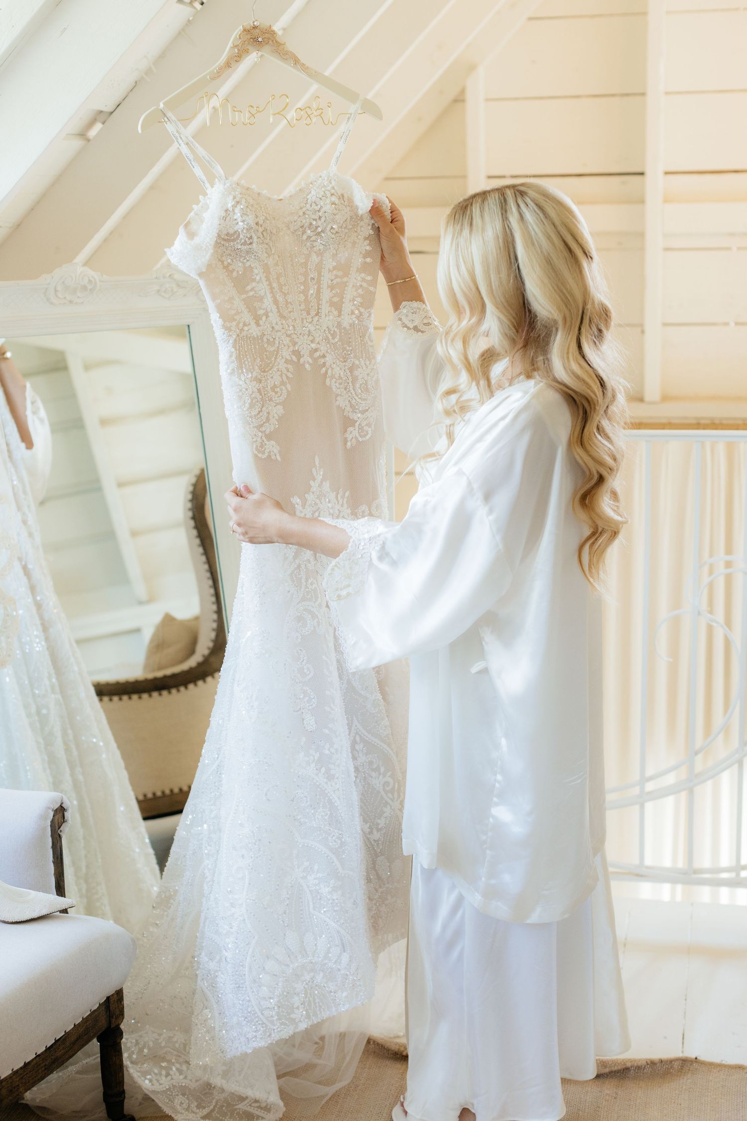 A beautiful white lace wedding dress hangs elegantly against a cream-colored wall.