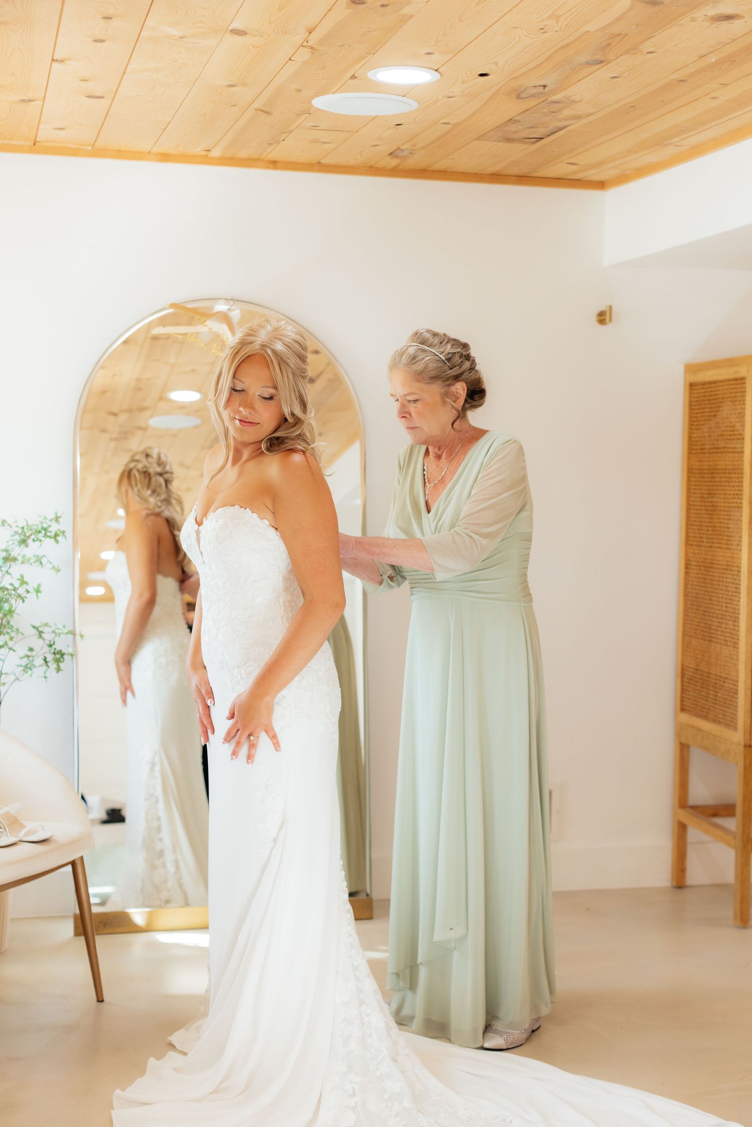 Bride getting final dress adjustments in bright room with mint green bridesmaid dress visible.