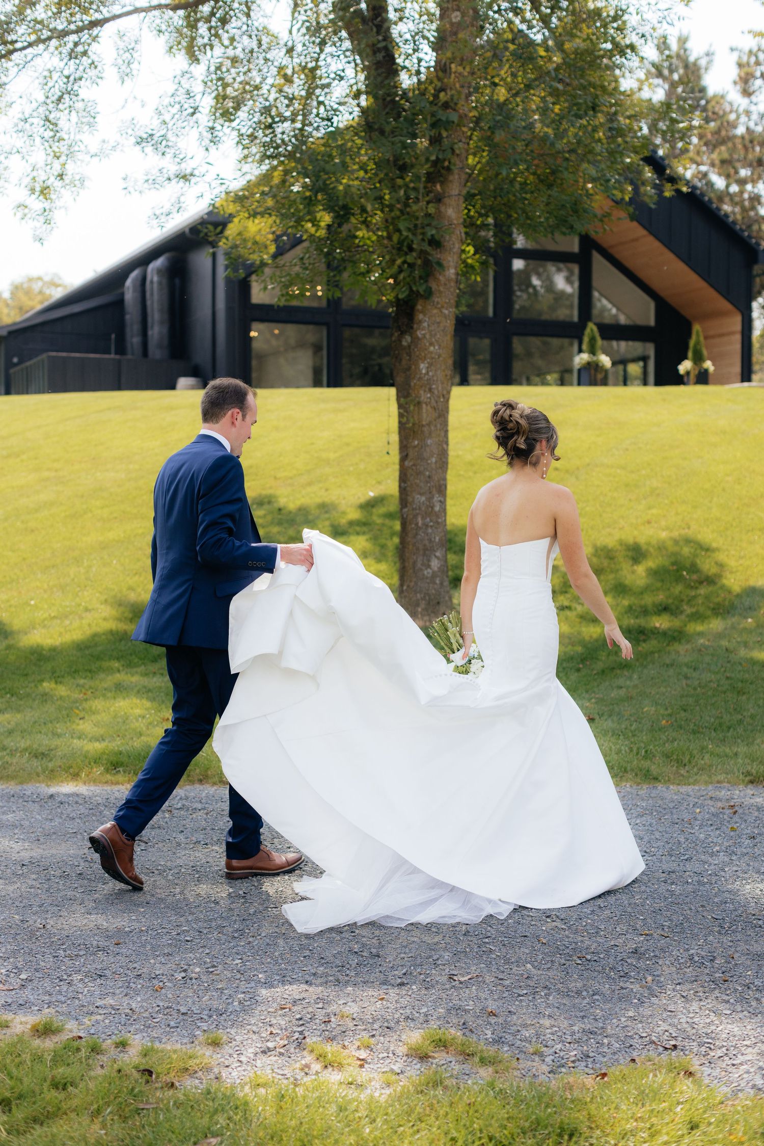 Wedding couple playfully walks along gravel path with dress trailing behind.