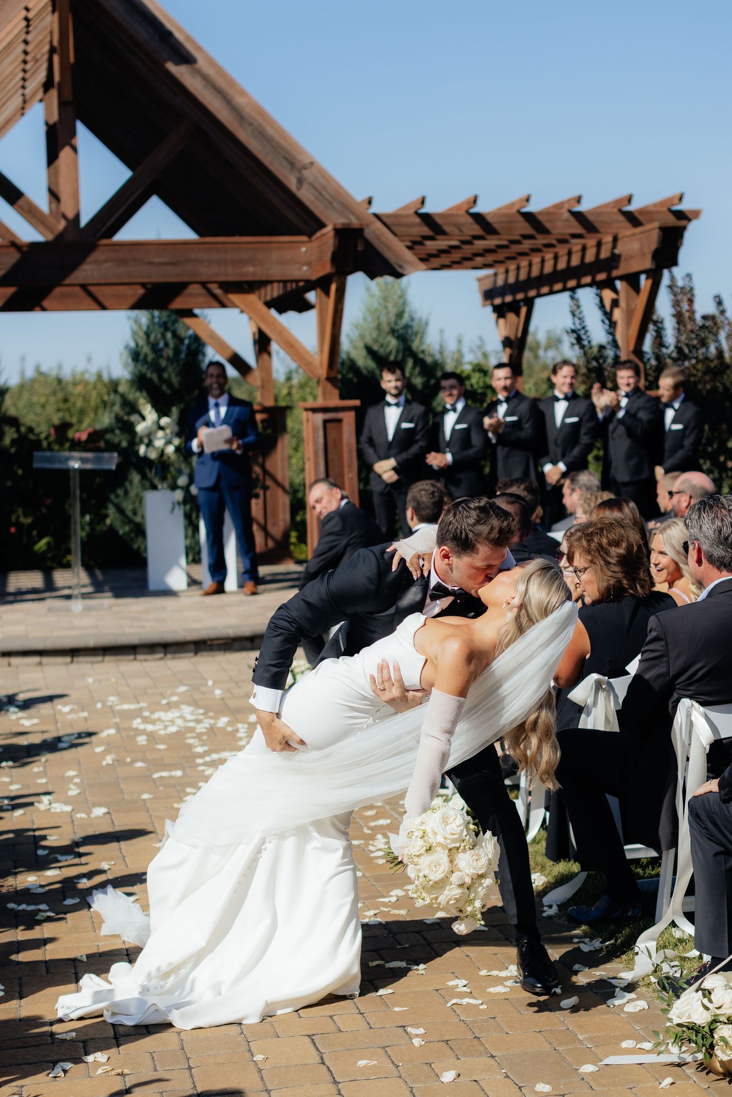 A dramatic dip kiss during an outdoor wedding ceremony under a wooden pergola with scattered petals on the ground.