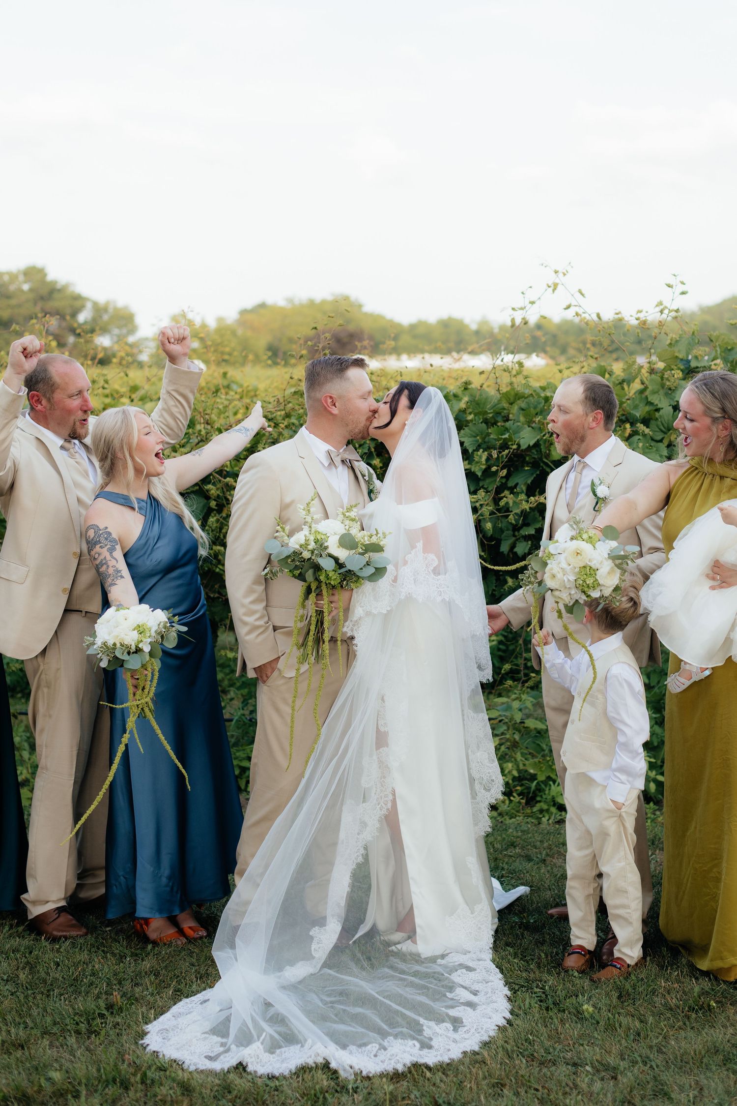 Wedding party in mix of tan suits and blue dresses poses together outdoors with bride and groom.