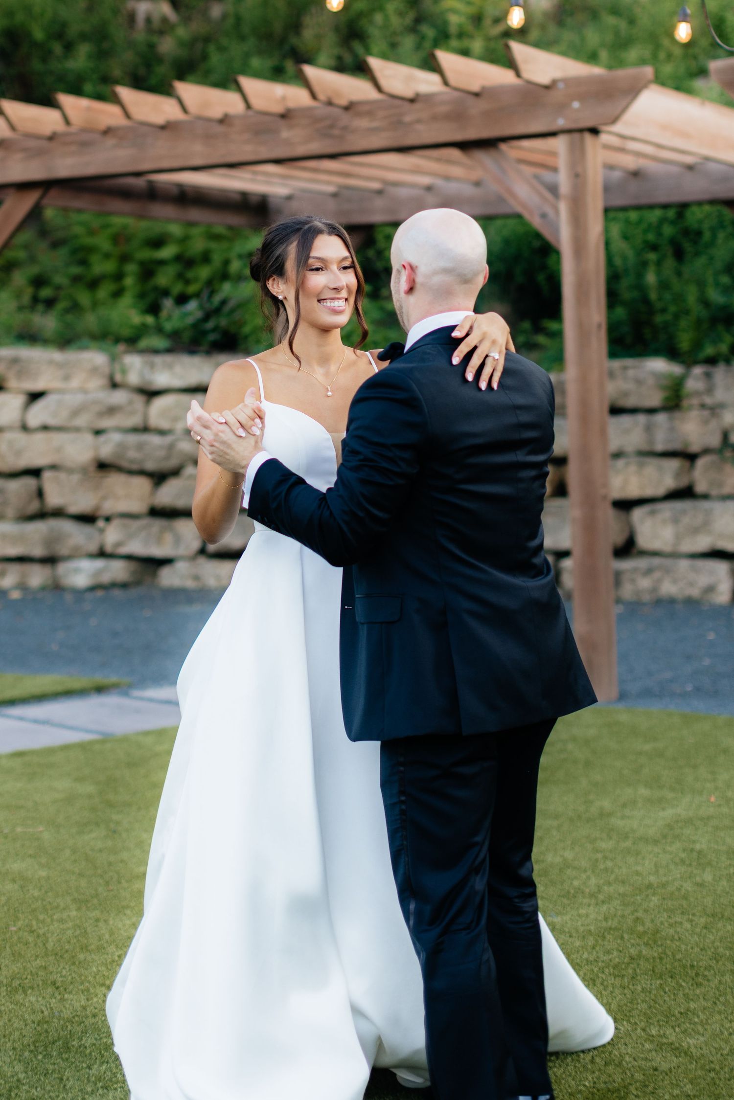 Newlyweds share a playful moment on grass under a wooden pergola.