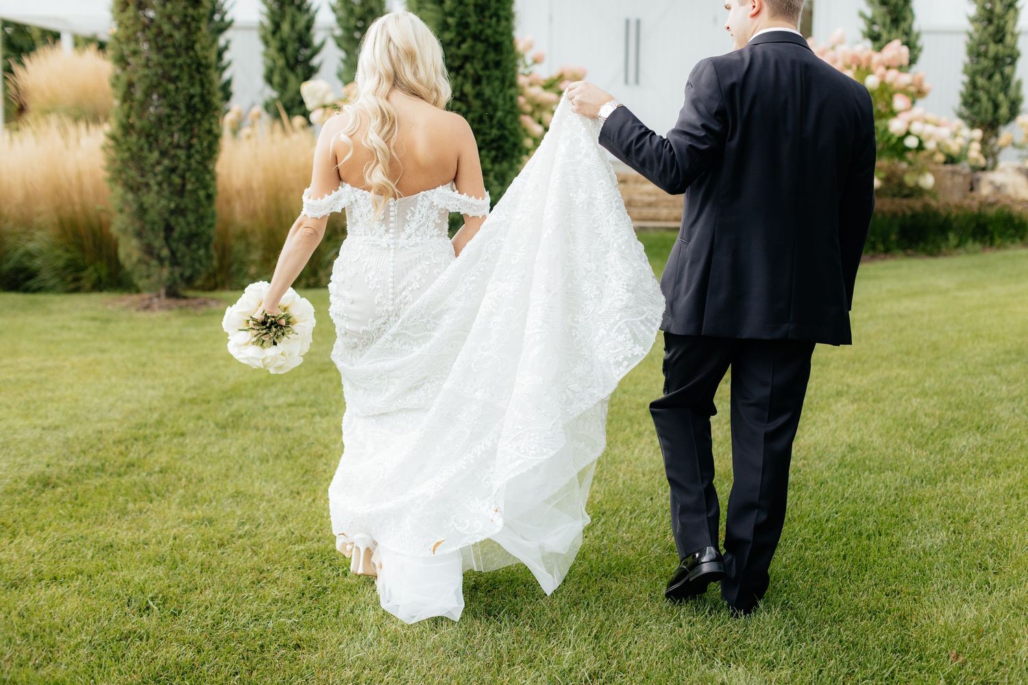 Bride and groom walk hand in hand across a lush green lawn during their outdoor wedding celebration.