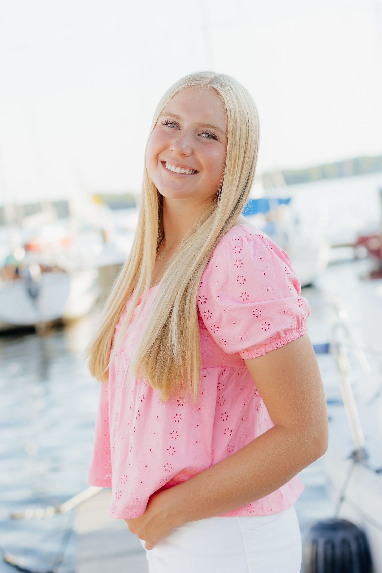 Woman in pink eyelet blouse smiling near a marina on a bright sunny day.