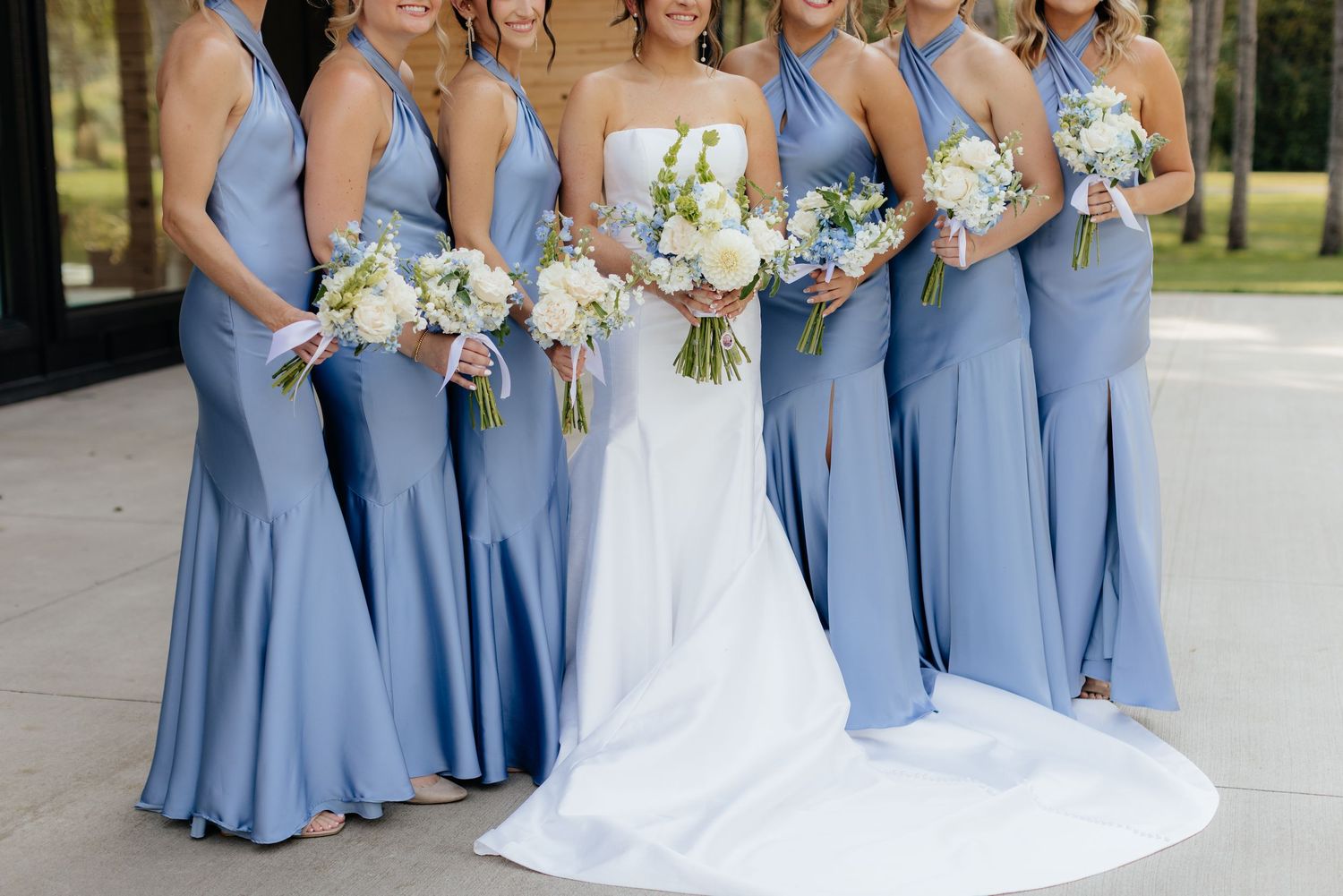 Wedding party in blue bridesmaid dresses pose with white floral bouquets.
